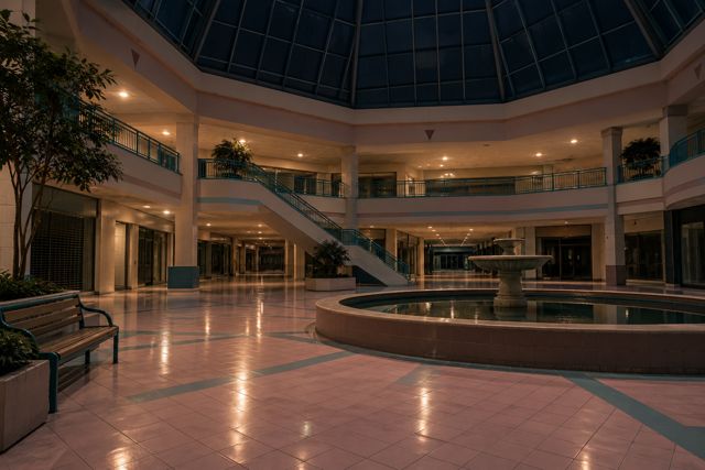 A pastel shopping-mall atrium sits empty beneath a skylight, with still water and an abandoned bench.
