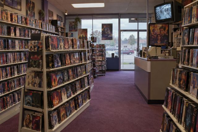 Shelves of tapes fill a small neighbourhood video shop, lit by a cloudy afternoon outside.