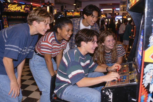 Friends crowd around a glowing arcade cabinet, with oversized denim, checkerboard floors, and the last coins of the evening.